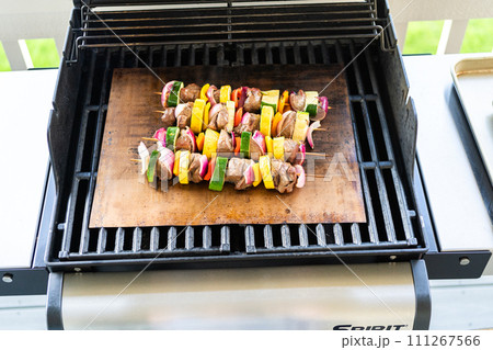 Skewered beef and fresh veggies sizzle on a copper grill mat over a gas outdoor grill. Skewered beef and fresh veggies sizzle on a copper grill mat over a gas outdoor grill. 111267566