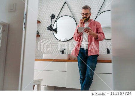 Man shopping online while brushing his teeth in bathroom. Holding tablet, scrolling and buying in eshop, toothbrush in mouth. 111267724