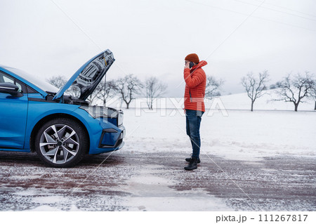 Angry man standing by electric car, battery run out of power before reaching destination. Man phone calling for help looking undre the car hood. Angry man standing by electric car, battery run out of power before reaching destination. Man phone calling for help looking undre the car hood. 111267817