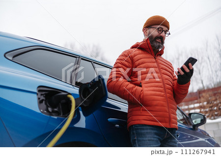 Man charging electric car during cold snowy day, using electric vehicle charging app, checking battery life, energy consumption on smart phone. Charging and driving electric vehicles during winter 111267941