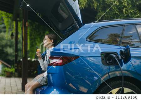 Girl and electric car with charger in charging port. Girl sitting in car trunk, eating apple while car is charging before going to school. 111269160