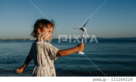 Small girl with blonde hair standing at beach, holding wind turbine model in hand, sea wind spinning blades. Small girl with blonde hair standing at beach, holding wind turbine model in hand, sea wind spinning blades. 111269229