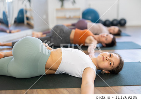 Sporty women doing yoga with group in studio, lying on mat with knee pressed to chest with hands, performing hamstring and low back stretching Ardha Pawanmuktasana 111269288