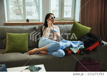Happy, joyful young woman sitting at home, holding plane tickets, talking on phone and expressing excitement about upcoming trip Happy, joyful young woman sitting at home, holding plane tickets, talking on phone and expressing excitement about upcoming trip 111271761