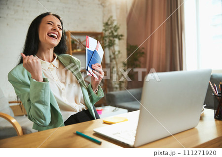 Happy, joyful young woman, employee sitting in office with laptop, holding plane tickets and showing excitement about upcoming trip Happy, joyful young woman, employee sitting in office with laptop, holding plane tickets and showing excitement about upcoming trip 111271920