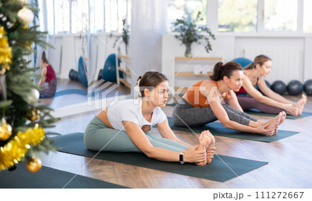 Active woman in sportswear with group of active people sitting on yoga mat and doing stretching legs during yoga class in fitness center during Christmas holidays Active woman in sportswear with group of active people sitting on yoga mat and doing stretching legs during yoga class in fitness center during Christmas holidays 111272667