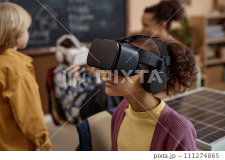 Side view portrait of young Black girl wearing VR headset in class and enjoying immersive learning experience copy space 111274865