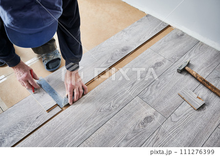 Man preparing laminate plank for floor installation in apartment under renovation. Close up of male construction worker using metal ruler and pencil while drawing line on laminate flooring board. Man preparing laminate plank for floor installation in apartment under renovation. Close up of male construction worker using metal ruler and pencil while drawing line on laminate flooring board. 111276399
