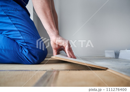 Male hands laying laminate wood plank on floor underlayment. Close up of man construction worker installing laminate timber flooring in apartment under renovation. Hardwood floor renovation concept. Male hands laying laminate wood plank on floor underlayment. Close up of man construction worker installing laminate timber flooring in apartment under renovation. Hardwood floor renovation concept. 111276400