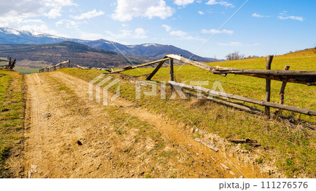 country road through the hill with grassy meadow behind the wooden fence. carpathian mountain ridge with snow capped tops in the distance. beauty of ukrainian countryside in spring country road through the hill with grassy meadow behind the wooden fence. carpathian mountain ridge with snow capped tops in the distance. beauty of ukrainian countryside in spring 111276576