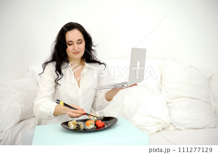 Pleasant caucasian lady sit on bed eating sushi while watching movie at home. Adorable short haired female in domestic wear have rest at weekends, holidays. leisure, relaxation, lifestyle 111277129