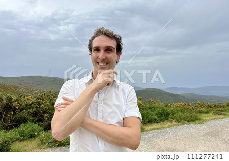 a handsome man of European appearance stands against the backdrop of mountains, he laughs a little touches his face with his hand 111277241