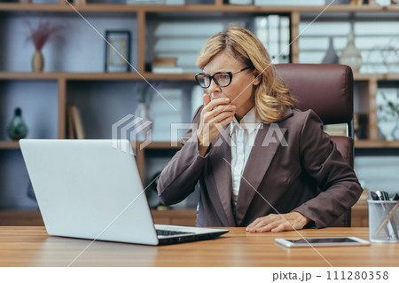 Shocked senior business woman sitting in the office at the desk and looking worriedly at the laptop screen. Problems with accounts, received bad news in the mail, reads the message. 111280358