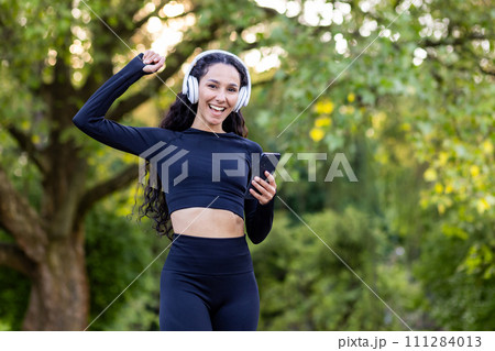 Happy woman in black activewear and white headphones raising hand with clenched fist while using smartphone. Sporty female with curly hair reaching goal of running marathon on public park background. Happy woman in black activewear and white headphones raising hand with clenched fist while using smartphone. Sporty female with curly hair reaching goal of running marathon on public park background. 111284013