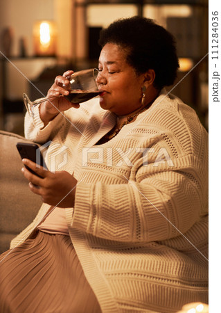 Vertical side view portrait of Black senior woman using smartphone and drinking wine during evening at home 111284036