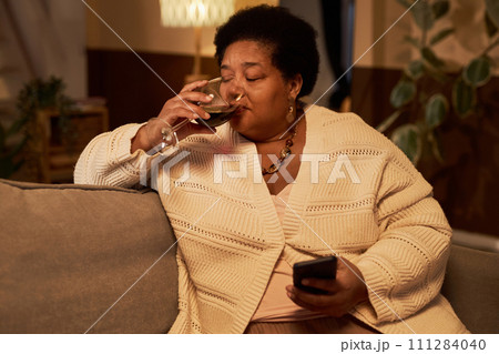 Portrait of African American senior woman using smartphone and drinking wine while enjoying relaxing evening at home, copy space Portrait of African American senior woman using smartphone and drinking wine while enjoying relaxing evening at home, copy space 111284040