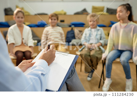 Close up of teacher or therapist holding clipboard and taking notes while working with group of childrenwith copy space 111284230