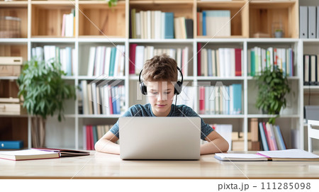 A focused young boy with headphones using a laptop at a desk with books, in a bright room with a bookshelf 111285098