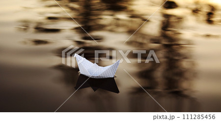 A paper boat is floating on the waves in the water at a beautiful sunset. Origami ship Sailing. The concept of a dream, future, childhood, freedom or hope. A paper boat is floating on the waves in the water at a beautiful sunset. Origami ship Sailing. The concept of a dream, future, childhood, freedom or hope. 111285456