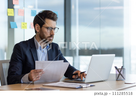 Serious and focused financier accountant on paper work inside office, mature man using calculator and laptop for calculating reports and summarizing accounts, businessman at work in suit. 111285518