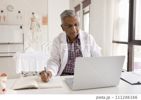 Mature Indian doctor man in uniform coat working at laptop Mature Indian doctor man in uniform coat working at laptop 111286166