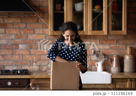 Excited woman unpacking carton box in kitchen, looking inside Excited woman unpacking carton box in kitchen, looking inside 111286169