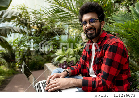 Hindu man in checkered shirt working on laptop in the garden Hindu man in checkered shirt working on laptop in the garden 111286673
