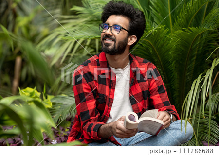 Hindu young man sitting in the garden with a book in hands Hindu young man sitting in the garden with a book in hands 111286688