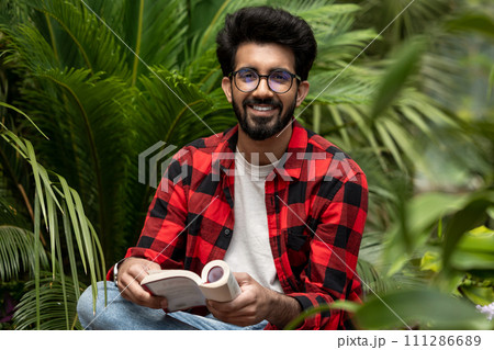 Hindu young man sitting in the garden with a book in hands Hindu young man sitting in the garden with a book in hands 111286689