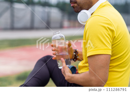 sportive man in a vibrant t-shirt, headphones, sits with a water bottle at the sports arena, taking a break from training. 111286791