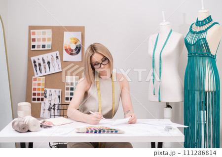 Thoughtful woman with glasses studies clothes sketch at desk in fashion atelier green macrame dress on mannequin near designer workplace in sewing shop Thoughtful woman with glasses studies clothes sketch at desk in fashion atelier green macrame dress on mannequin near designer workplace in sewing shop 111286814