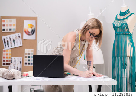 Woman designer marks notes at desk with laptop in dressmaking workshop tailor creates women clothes by mannequin with macrame dress in fashion atelier 111286815