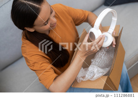 Lady sits on couch in living room of accommodation unpacking cardboard box with light headphones smiling female with dark hair opens unwrap carton parcel at home Lady sits on couch in living room of accommodation unpacking cardboard box with light headphones smiling female with dark hair opens unwrap carton parcel at home 111286881