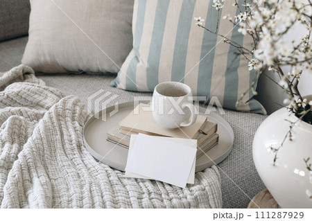 Spring breakfast scene. Blank greeting card mockup. Cup of coffee, tea on books. Round beige tray. Blossoming cherry plum tree branches in ceramic vase. Cozy linen sofa, cushions. Easter still life. Spring breakfast scene. Blank greeting card mockup. Cup of coffee, tea on books. Round beige tray. Blossoming cherry plum tree branches in ceramic vase. Cozy linen sofa, cushions. Easter still life. 111287929