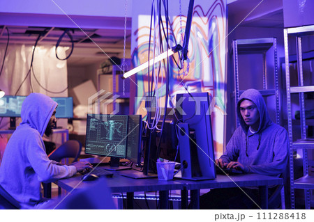 Hackers collaborating while coding virus software, doing illegal activities and breaking law in abandoned warehouse. Criminals programming malware on computers in hideout place Hackers collaborating while coding virus software, doing illegal activities and breaking law in abandoned warehouse. Criminals programming malware on computers in hideout place 111288418