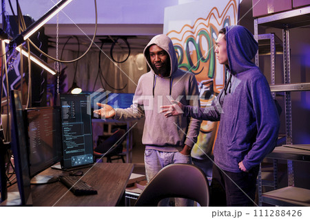 Hackers discussing hacking plan and cybercrime technique. Criminals in hood standing near computer and talking about malicious software code on screen in abandoned warehouse Hackers discussing hacking plan and cybercrime technique. Criminals in hood standing near computer and talking about malicious software code on screen in abandoned warehouse 111288426