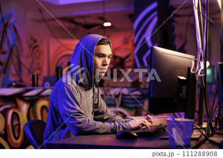 Asian internet scammer cracking password on computer to get access to database. Young man in hood writing malicious software code on computer and breaking into server in dark room Asian internet scammer cracking password on computer to get access to database. Young man in hood writing malicious software code on computer and breaking into server in dark room 111288908