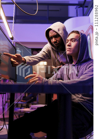 Confused hacker pointing at computer screen and asking criminal about password cracking technique. Diverse young men in hoods planning phishing attack while breaking law together Confused hacker pointing at computer screen and asking criminal about password cracking technique. Diverse young men in hoods planning phishing attack while breaking law together 111288962