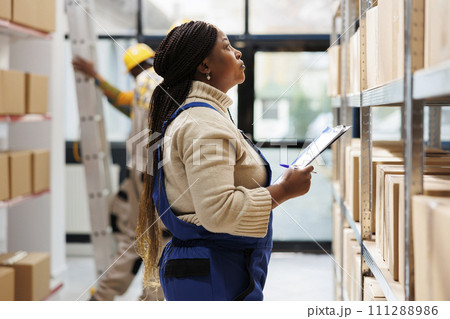 Warehouse supervisor looking at parcels and taking notes in storage room. African american logistics manager holding clipboard with order checklist and planning distribution 111288986