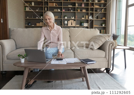 Positive mature woman pays bills online use calculator and laptop 111292605