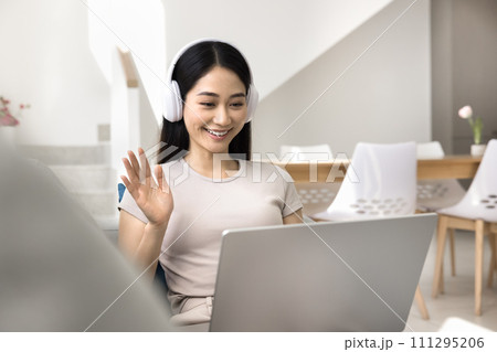Cheerful young Asian student girl in headphones waving hand hello Cheerful young Asian student girl in headphones waving hand hello 111295206