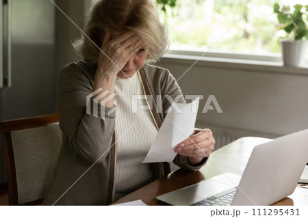 Confused older woman sit at desk look at overdue bill Confused older woman sit at desk look at overdue bill 111295431