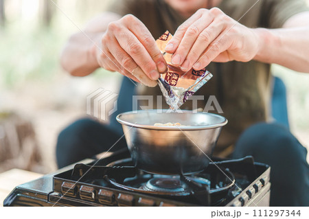 キャンプ飯でインスタントラーメン・袋麺に液体スープを入れるキャンパーの男性 111297344