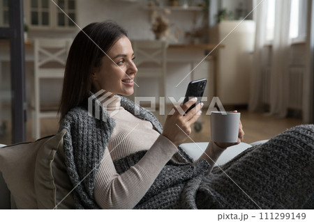 Woman covered with plaid hold teacup, smartphone relaxing at home 111299149