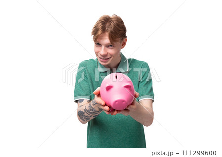 a young European man with red hair holds out a pink piggy bank with money and coins a young European man with red hair holds out a pink piggy bank with money and coins 111299600