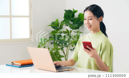 部屋でノートパソコンとスマホを使う女性 111300009