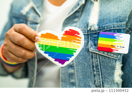 Asian lady wearing rainbow flag wristbands and hold red heart, symbol of LGBT pride month celebrate annual in June social of gay, lesbian, bisexual, transgender, human rights. 111300452