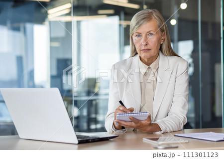 Concentrated senior woman looking at camera with focused sigh while holding notebook and pen by desktop with laptop. Experienced hr manager hosting interview and writing down features of applicant. 111301123