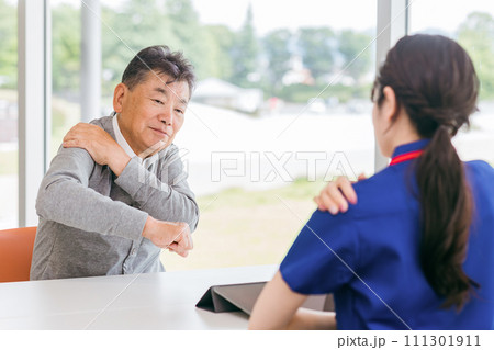 女性看護師・理学療法士に肩の痛みを相談するシニア男性(リハビリ・関節痛) 女性看護師・理学療法士に肩の痛みを相談するシニア男性(リハビリ・関節痛) 111301911
