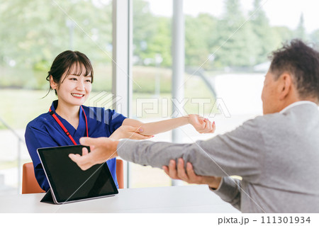 高齢者男性患者の肘の痛みの相談を聞く看護師・理学療法士の女性 111301934